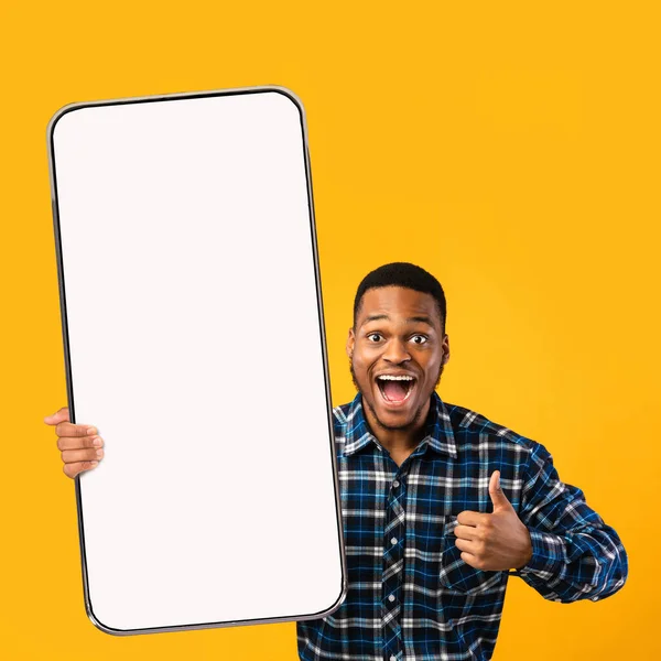 Cool aplicación móvil. Hombre negro lleno de alegría demostrando el teléfono celular con la pantalla vacía, mostrando el gesto hacia arriba pulgar, maqueta —  Fotos de Stock
