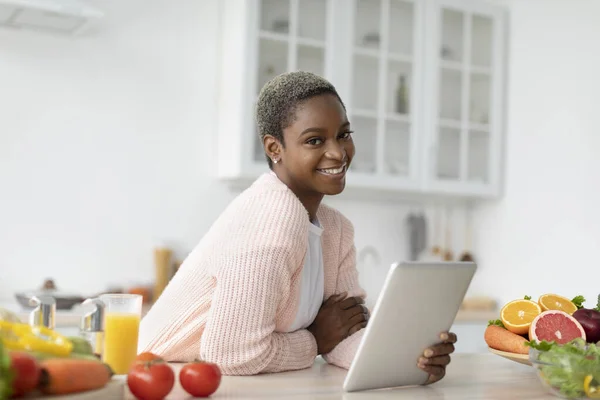 Glad millennial african american lady in casual with tablet in kitchen interior at table with vegetables — Stock Photo, Image