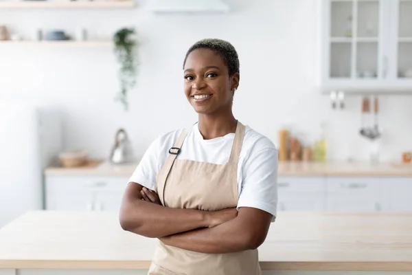 Cheerful millennial african american female in apron with crossed arms looking at camera at modern minimalist kitchen — Stock Photo, Image