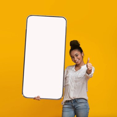 Büyük akıllı telefonu elinde tutan mutlu siyahi kadın boş ekranlı ve baş parmağını kaldırıyor, turuncu arka plan, model.