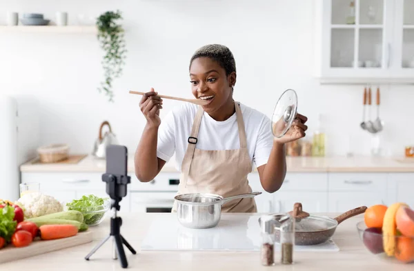 Glad millennial african american female in apron cooks at home and tasting dish shooting video lesson by phone — Stock Photo, Image