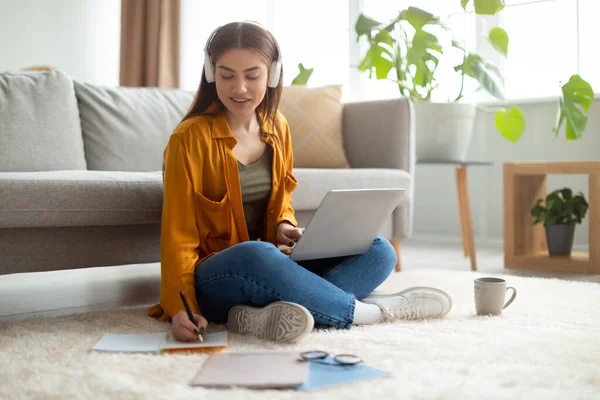 Mulher caucasiana feliz em fones de ouvido tendo reunião on-line no laptop, escrevendo informações durante o webinar em casa — Fotografia de Stock