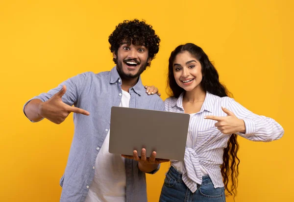 Ενθουσιασμένο ζευγάρι Ινδιάνων κρατώντας φορητό υπολογιστή και δείχνοντας τα δάχτυλα σε gadget μαζί, κίτρινο φόντο στούντιο — Φωτογραφία Αρχείου