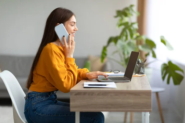 Mujer joven caucásica usando el ordenador portátil y hablando en el teléfono inteligente, trabajando en línea desde casa, espacio de copia — Foto de Stock