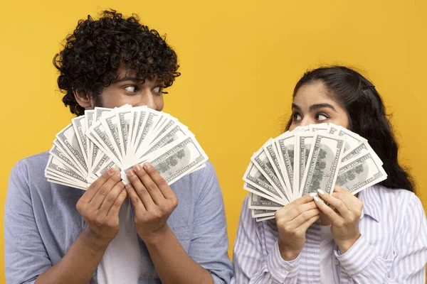 Gran concepto de ganar. Joven pareja india sosteniendo y cubriendo caras con un montón de dólares ventilador de efectivo, fondo de estudio amarillo — Foto de Stock