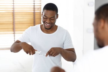 Diş fırçasına diş macunu süren yakışıklı siyah adam banyoda aynanın yanında.