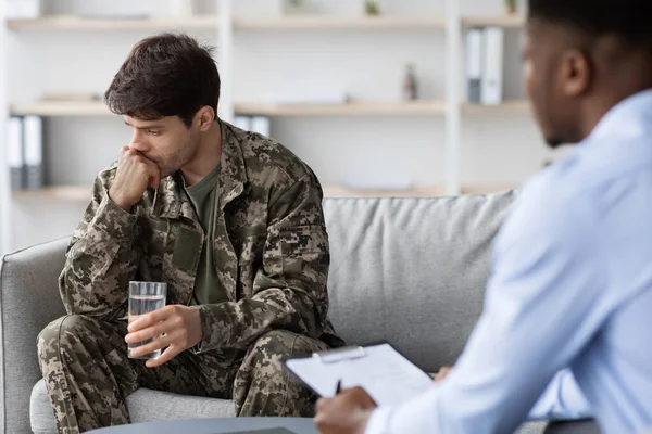 Thoughtful soldier at therapy with psychologist, holding glass of water