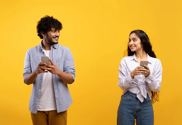 Joven indio alegre hombre y mujer en trajes casuales sosteniendo teléfonos inteligentes, mirándose y sonriendo — Foto de Stock