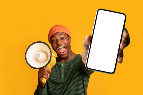 Emocionado cara negro gritando em megafone, mostrando smartphone grande com tela vazia, oferecendo espaço mockup para anúncio — Fotografia de Stock
