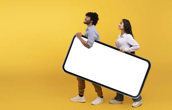 Mladý indický pár drží obří smartphone s mockup pro design, žluté pozadí studia, volné místo — Stock fotografie