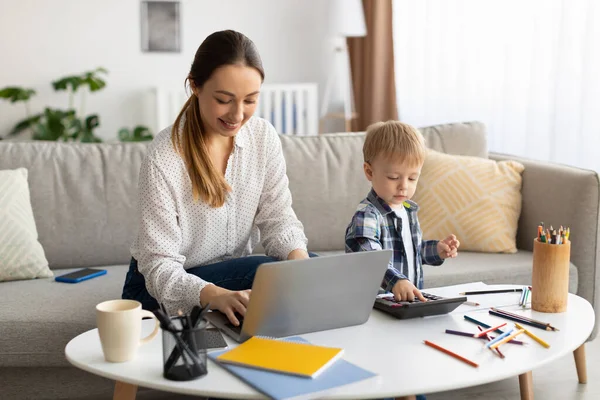 Фрилансер в декретном отпуске. Молодая мать работает на ноутбуке, в то время как ее маленький сын играет с калькулятором neaby — стоковое фото