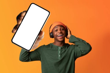 Kulaklıklı heyecanlı siyah adam boş ekranlı büyük bir cep telefonu gösteriyor. Model için yeri olan bir telefon gösteriyor.