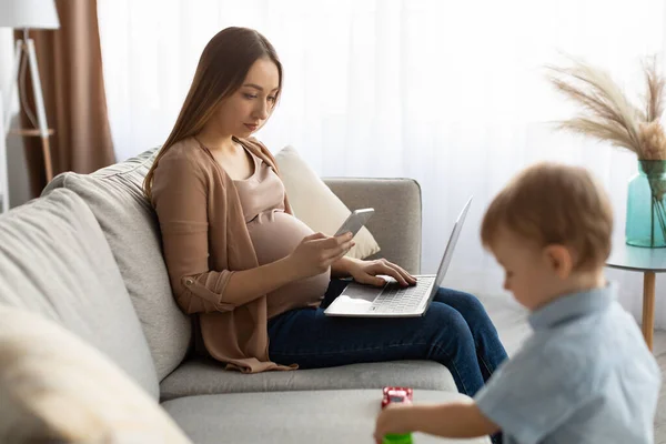 Jeune femme indépendante enceinte utilisant un smartphone et un ordinateur portable, maman prenant soin de son fils tout-petit — Photo