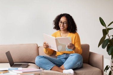 Neşeli Afrikalı Amerikalı kadın dizüstü bilgisayarı ve kitabı ile koltukta oturuyor, ev ödevi yapıyor, evde kitap okuyor, boş yer arıyor.