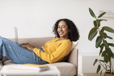 Neşeli genç siyahi kadın dizüstü bilgisayarla iletişim kuruyor oturma odasında kanepede oturuyor.