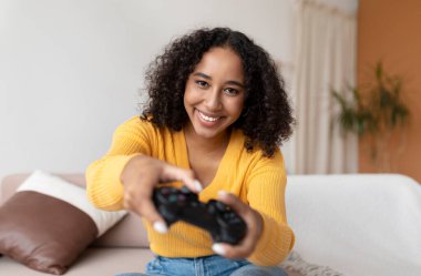 Pozitif genç siyahi kadın joystick kullanıyor, evdeki kanepede video oyunu oynuyor.