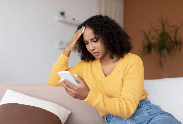 Molesto joven mujer negra leyendo malas noticias o sms en el teléfono celular, sentado en el sofá en casa —  Fotos de Stock