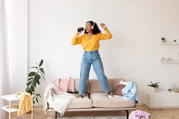 Cool femme noire dans les écouteurs en utilisant le smartphone comme micro, debout sur le canapé, chantant des chansons, vérifier application de musique mobile — Photo