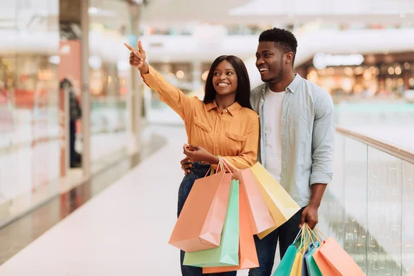 Cheerful black couple holding shopping bags pointing at window — Stock Photo, Image