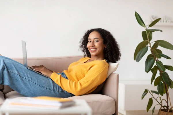 Alegre joven negro mujer comunicarse en línea a través de ordenador portátil, sentado en el sofá en la sala de estar —  Fotos de Stock