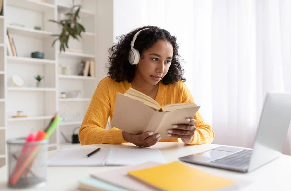 Portré fiatal fekete hölgy vezeték nélküli fejhallgató ül az asztalnál, a laptop és az olvasás könyv otthon — Stock Fotó