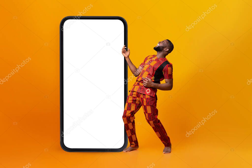 Joyful black man in traditional costume dancing near huge cellphone with blank white screen, mockup