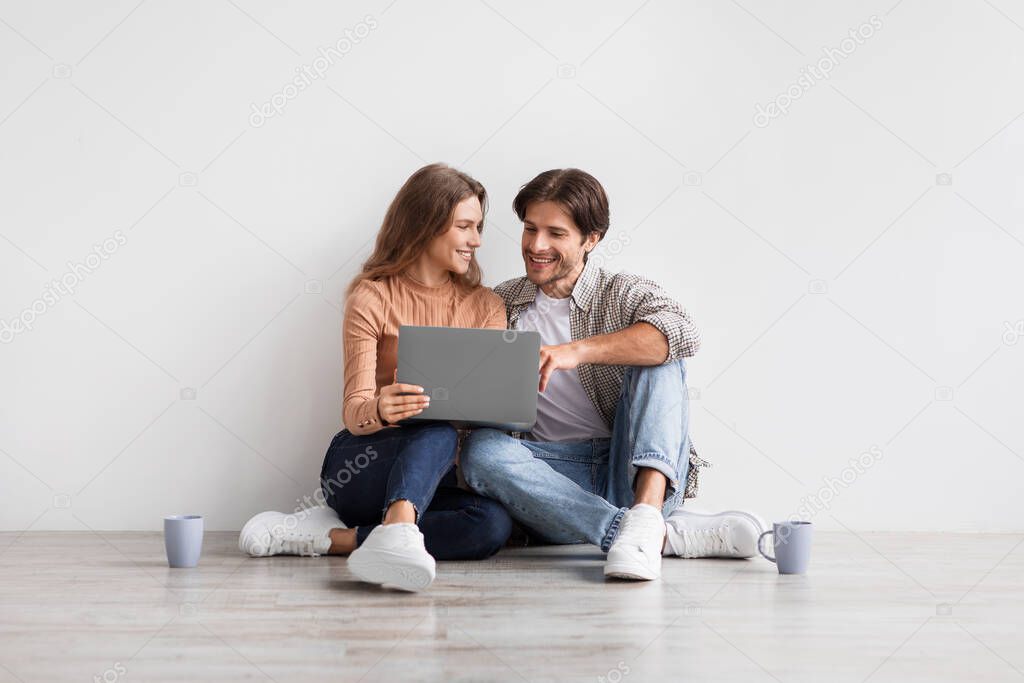 Glad young european man and lady planning future interior with laptop, sit on floor with coffee on white wall background
