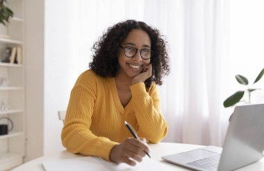 Mutlu genç siyah bayan evde dizüstü bilgisayarla çalışıyor, uzaktan ders sırasında notlar alıyor, uzaktan öğrenmenin keyfini çıkarıyor.