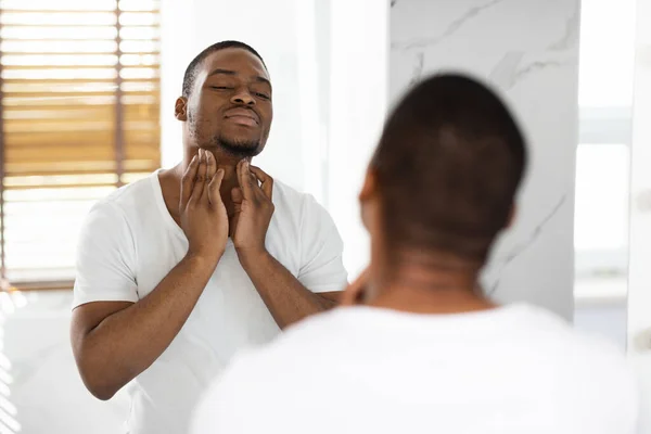 Concepto de Salud. Hombre negro preocupado tocando el cuello, examinando los ganglios linfáticos cerca del espejo — Foto de Stock