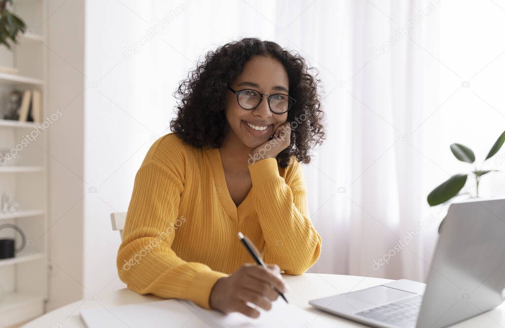 Happy young black lady studying online with laptop at home, taking notes during remote lecture, enjoying distance learning