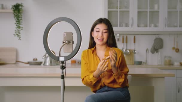 年轻的亚洲女性美容美发博主坐在厨房里，手持智能手机和圆形灯，记录着关于新的轮廓线的视频 — 图库视频影像