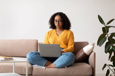 Kanepede bacak bacak üstüne atmış vaziyette oturan odaklanmış genç siyahi bir bayan evde online iş veya eğitim için dizüstü bilgisayar kullanıyor.