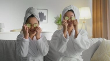 Mutlu Asyalı anne ve kızının portresini büyüt. Kafalarına havlular ve bornozlar giyip gözlerini salatalıkla kapatıyorlar.