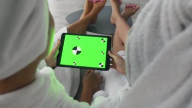 Tanımlanamayan anne ve kız bornoz ve havlu giyip tablette krom ekranlı dijital televizyon izliyorlar.