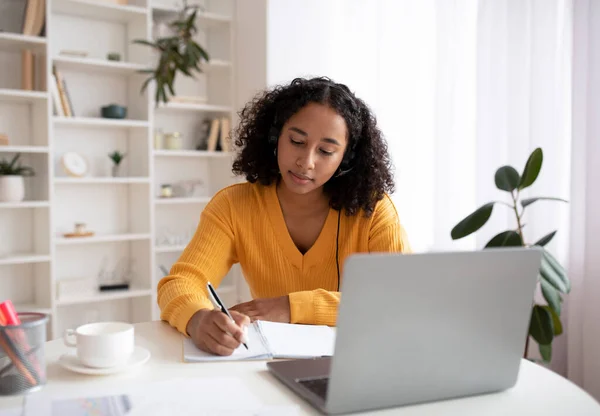 Cheery young black woman in headphones having online lesson on laptop, writing in notebook at home — Stock Photo, Image