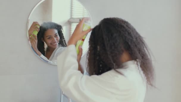 Молодая африканская американка наносит лак для волос на кудрявые волосы, делает прическу дома, смотрит в зеркало в ванной комнате — стоковое видео