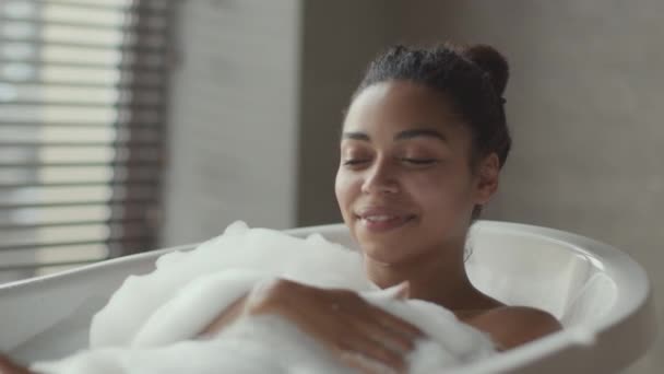 Badskoj. Ung bekymmerslös afrikansk amerikansk dam leker med skum i badkaret, koppla av i badkaret i badrummet, slow motion — Stockvideo