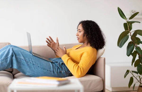 Jovem afro-americana positiva usando laptop, relaxando no sofá em casa, vista lateral — Fotografia de Stock