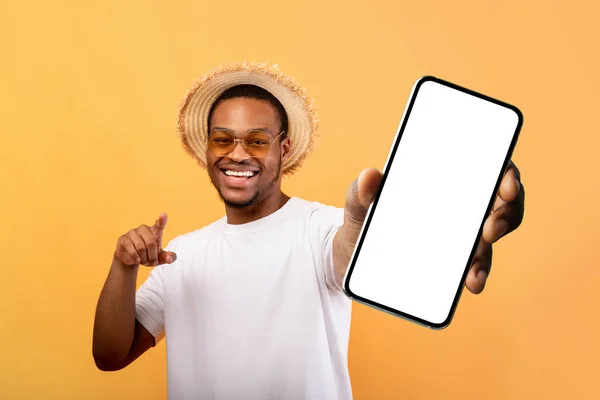 Podekscytowany czarny mężczyzna w letniej czapce pokazuje telefon komórkowy z pustym ekranem, polecając aplikację mobilną do podróży — Zdjęcie stockowe