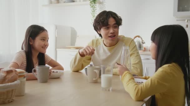 Szczęśliwej rodzinnej rozrywki. Młody pozytywny Azji ojciec rozmawia z małymi córkami, korzystając śniadanie razem w kuchni — Wideo stockowe