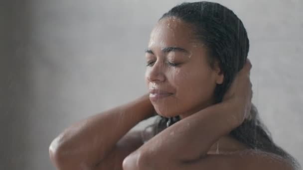 Concepto de higiene matutina. Joven mujer afroamericana tomando ducha, de pie bajo gotas de agua en el baño — Vídeo de stock