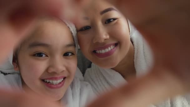 Uśmiechnięta azjatycka matka i córka w białych szlafrokach z symbolem serca, łączącymi dłonie — Wideo stockowe