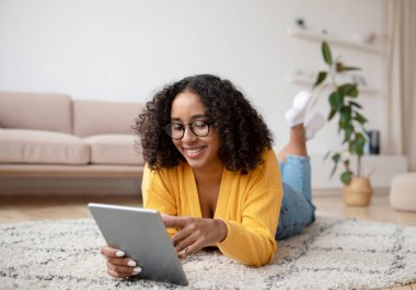 Siyah kadın evde yerde yatıyor, dijital tablette yeni mobil uygulamalar kullanıyor, internetten alışveriş yapıyor, arkadaşıyla görüntülü sohbet ediyor.