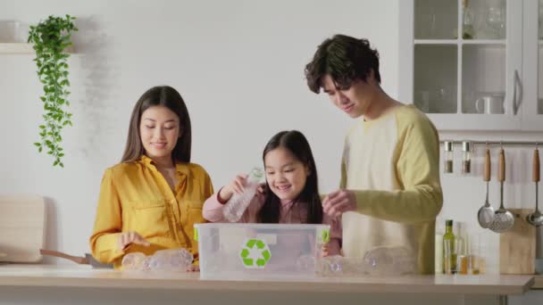 Jóvenes padres asiáticos enseñando a su hija a un ambiente seguro, recogiendo botellas de plástico para reciclar en la cocina — Vídeos de Stock