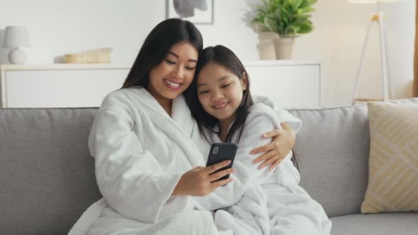 Genç kaygısız Asiana anne internette akıllı telefondan sörf yapıyor, küçük kızıyla kanepede kucaklaşıyor. — Stok video