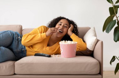 Genç siyahi kadın kanepede dinleniyor, TV izliyor ve evde patlamış mısır yiyor. Tembel Afro-Amerikan kadın televizyon programından ya da filmden zevk alıyor, oturma odasında atıştırıyor.
