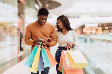 Perakende ve Teknoloji. Akıllı telefon kullanan mutlu Afro-Amerikan çiftin portresi, alet ekranını gösteren kadın, cep telefonu ve alışveriş torbası tutan adam, şehir merkezinde yürüyen eşler.