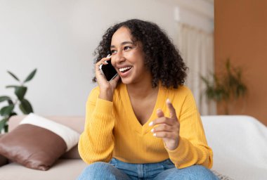 Akıllı telefondan konuşan, evdeki kanepede oturan pozitif genç siyahi bir kadın. Milenyum Afrikalı Amerikalı kadın oturma odasında cep telefonuyla konuşuyor.