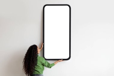 Beyaz duvarda reklamınız için boş ekranı olan kocaman akıllı telefonu olan bir kadın. Uygulama ya da web sitesi tasarımınız için bir model