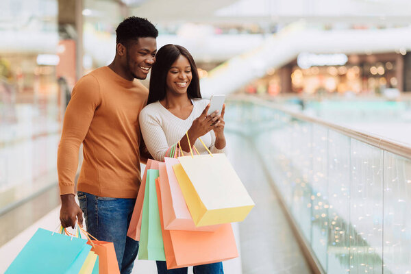 Черные покупатели совершают покупки с помощью мобильных телефонов, держа в руках разноцветные сумки для покупок, стоящие в холле, свободном пространстве для копирования. Счастливые клиенты с помощью приложения покупают одежду онлайн Via. Концепция электронной коммерции